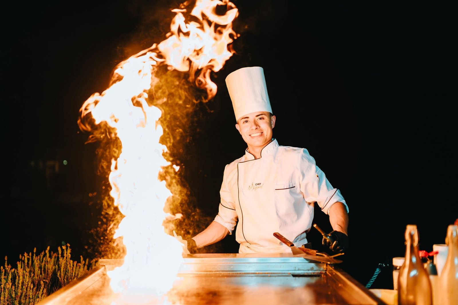 Chef Miguel smiling behind the teppanyaki grill with a tall flame rising beside him
